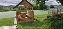 Wooden gazebo with a table and benches on a grassy area with a sports field in the background.